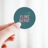 Sticker ‘Fijne kerst’ in petrol en roze tinten, vastgehouden in de hand